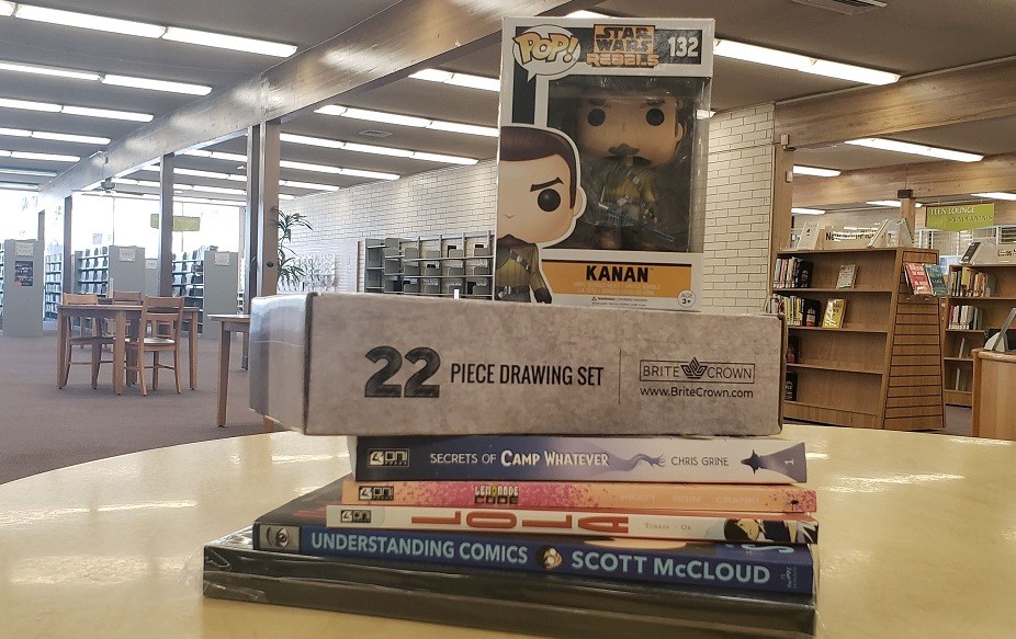Photo of a stack of prizes, including: a Funko Pop , 22 piece drawing set, a drawing journal, and four books: Secreta of Camp Whatever, Lola, Lemonade Code, and Understanding Comics.